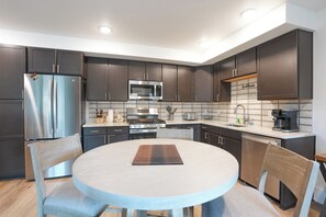 A modern kitchen with dark cabinets, stainless steel appliances, a round dining table, and a breakfast bar. - Blue River Flats Basecamp Condo: Walk to Blue Bird Market & Shops (Silverthorne)