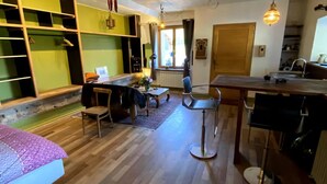 Un salon chaleureux avec un plancher en bois, un coin repas avec une table et des chaises, une cuisine équipée d’un évier et une grande fenêtre laissant entrer la lumière naturelle.