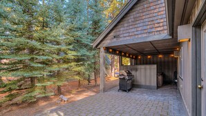 Une terrasse extérieure couverte, dotée d’un barbecue, entourée d’arbres et bordée d’un bâtiment en bois.