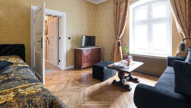 A room with a herringbone patterned floor, a bed with a patterned duvet, a dark sofa, a small table with a vase, a TV on a wooden cabinet, and a window with curtains. - Malila Apartments Cracow Lubicz 14 (Kraków)