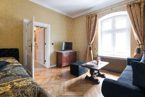 Une pièce avec un parquet à motifs en chevrons, un lit recouvert d’une couette à motifs, un canapé foncé, une petite table avec un vase, un téléviseur posé sur un meuble en bois et une fenêtre avec des rideaux.