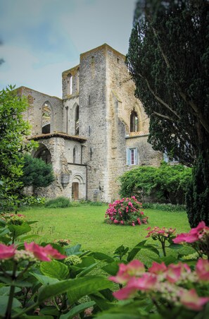 Ein historisches Steinhaus mit Rundbogenfenstern und einer mittleren Tür, umgeben von üppiger Vegetation und blühenden Pflanzen.