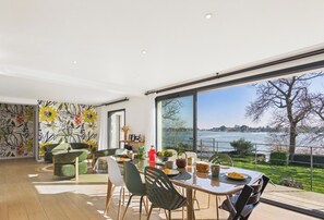 A dining area with a view of a lake, a large window, and a floral wall. - -FLORA- Family home on Penfoulic cove (La Forêt-Fouesnant)