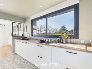A modern kitchen with a large window, white cabinetry, and a countertop with various kitchen items. - -FLORA- Family home on Penfoulic cove (La Forêt-Fouesnant)