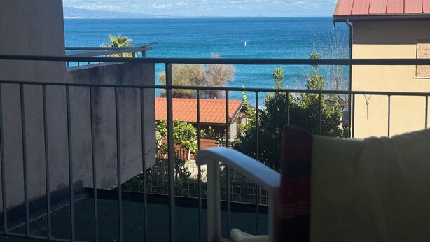 Un balcó amb vista al mar, una cadira i un edifici amb teulada vermella.