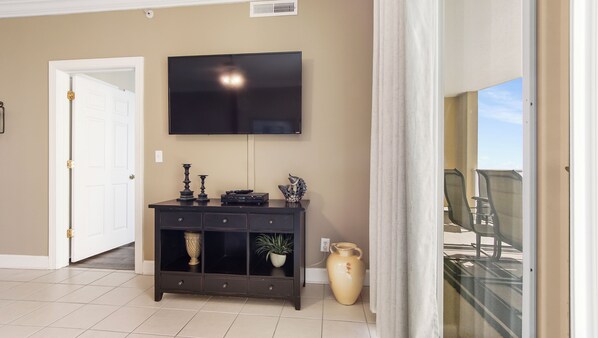A living room with a flat-screen TV mounted on the wall, a dark wooden console table with decorative items, a potted plant, and a large vase. - Emerald Isle 1808 - Front row chair service included! Affordable rates! (Panama City Beach)