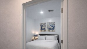 Un dormitorio con una cama, una mesita de noche, dos cuadros en la pared y una rejilla de ventilación en el techo.