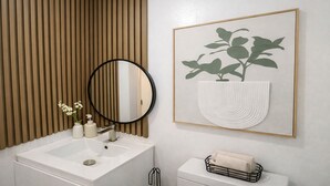 Un baño con lavamanos blanco, espejo redondo, obra de arte con planta enmarco y retrete con toallero.