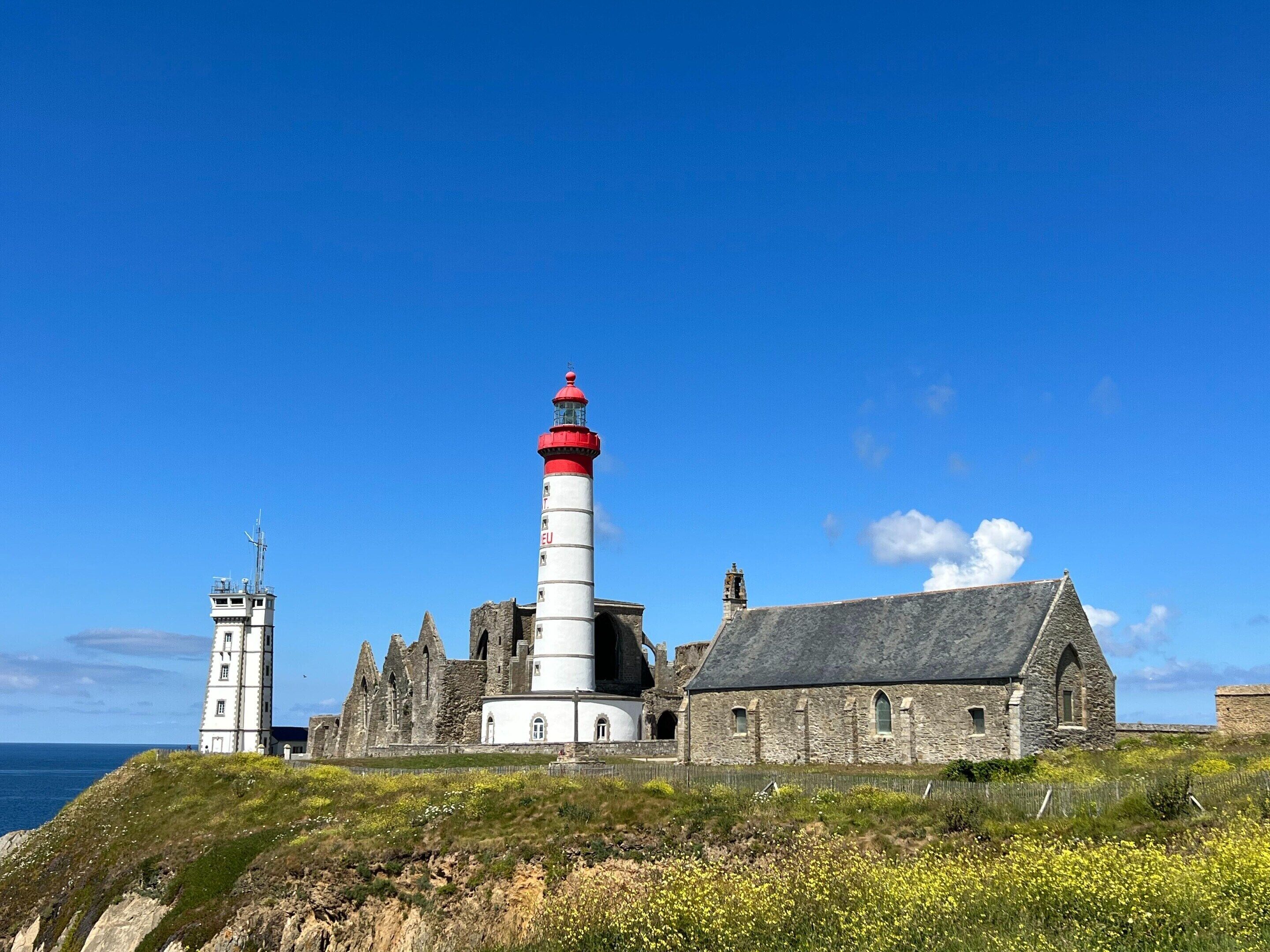 A coastal lighthouse with a red top, adjacent stone buildings, and a clear blue sky.