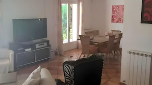 A living room with a TV, a dining area with chairs, a radiator, and a window with curtains. - Villa With Private Pool, Garden & Panoramic Views (Draguignan)