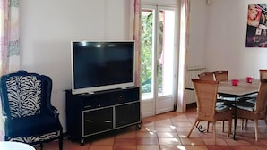 A living room with a TV on a stand, a dining table with chairs, and a patterned armchair. - Villa With Private Pool, Garden & Panoramic Views (Draguignan)