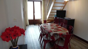 A dining area with a table covered in a patchwork tablecloth, surrounded by red chairs. A vase with red flowers is on the table, and a television is on a cabinet to the right. - Calme et détente assurée (Avèze)