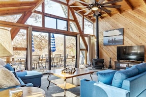 Un intérieur de chalet chaleureux avec un grand plafond en bois, une cheminée, un téléviseur et un coin repas offrant une vue sur l’extérieur.