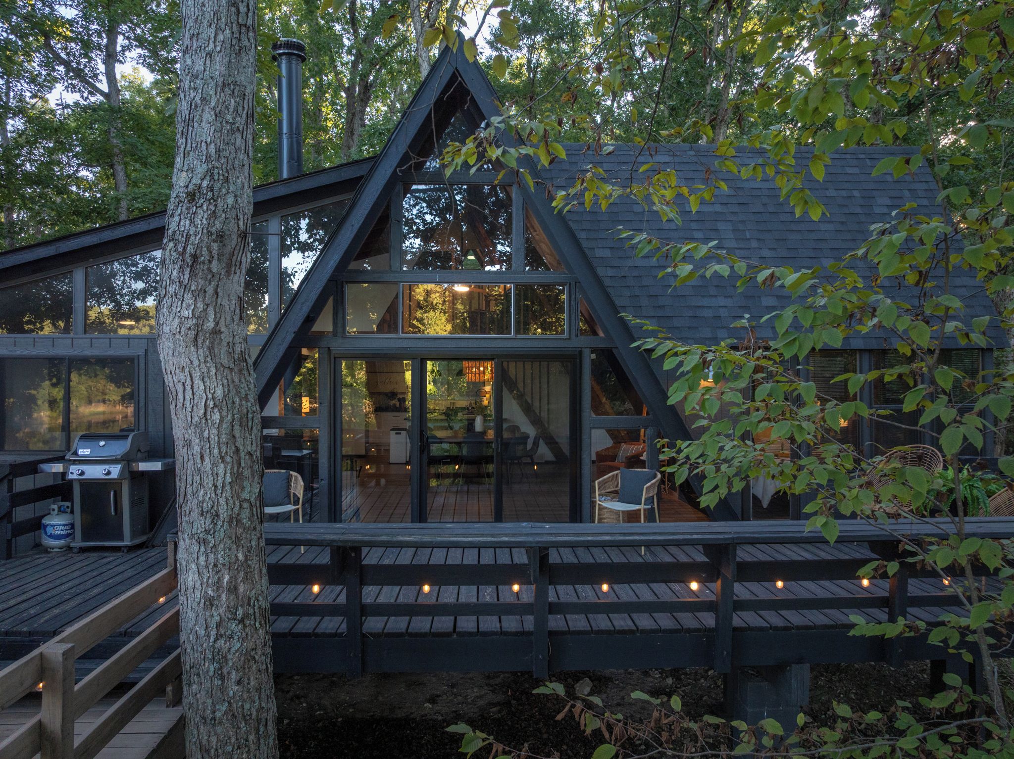A cabin with a deck, a grill, and a large window overlooking a forest.