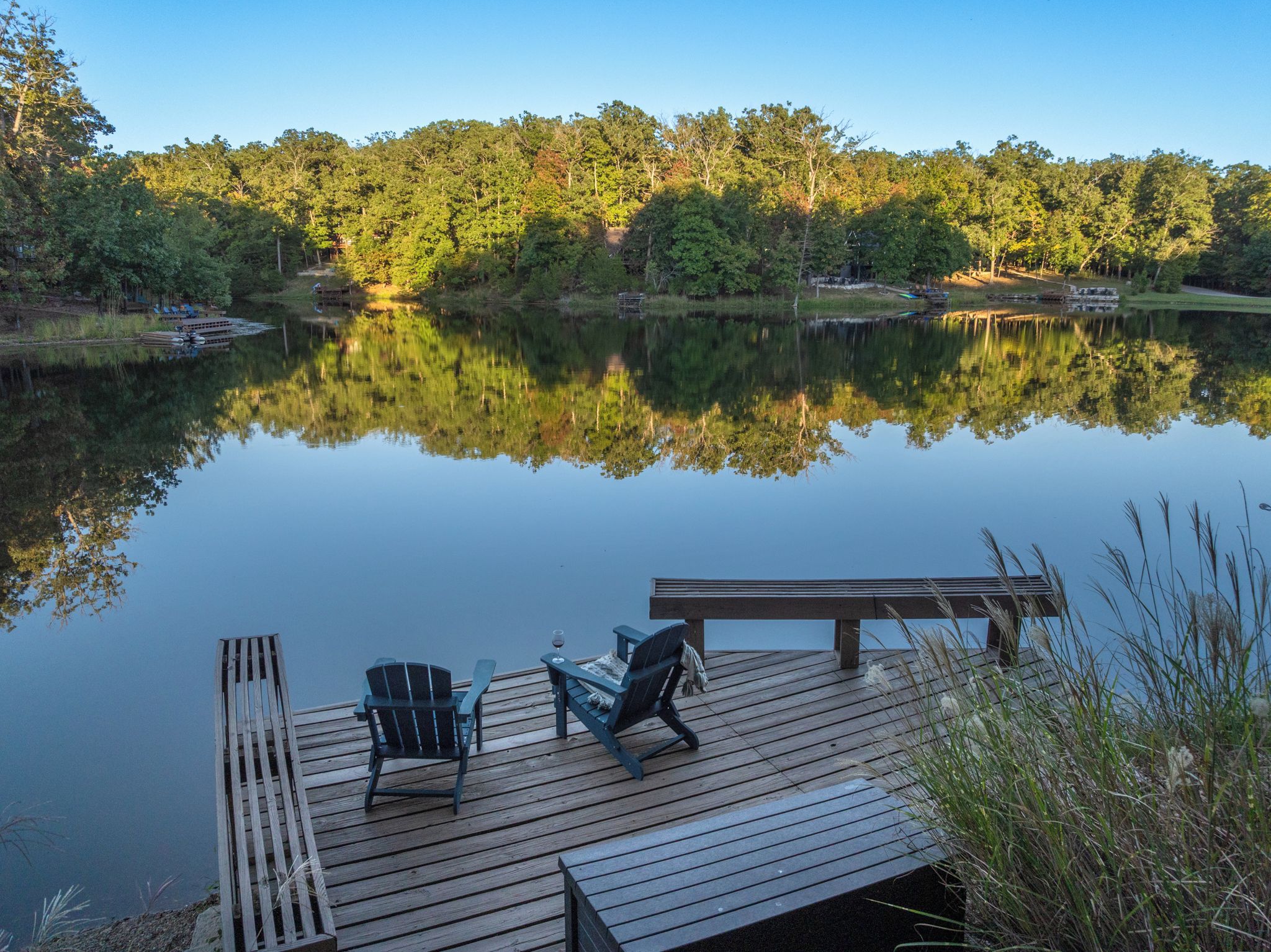 A calm lake with a wooden dock, two chairs, and a bench.