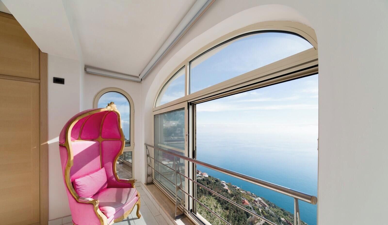 Ein Balkon mit einem rosa Stuhl und einem großen Fenster mit Blick auf die Küste.
