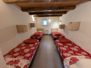 Een smalle kamer met vier eenpersoonsbedden op een rij, elk met een rode bedspreide en een houten hoofdbord. Aan weerszijden van de kamer bevinden zich wandlampen, en aan het uiteinde een klein raam met een ronde ruit.