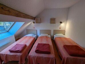 A room with three single beds, each with a pink bedspread and a folded towel. There are two wall-mounted light fixtures and a small window with a view of trees.