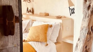 Ein Schlafzimmer mit Holzboden, einem Bett mit weißer und senffarbener Bettwäsche, einem Nachttisch mit Lampe sowie einem an der Wand befestigten Regal mit Dekorationen.