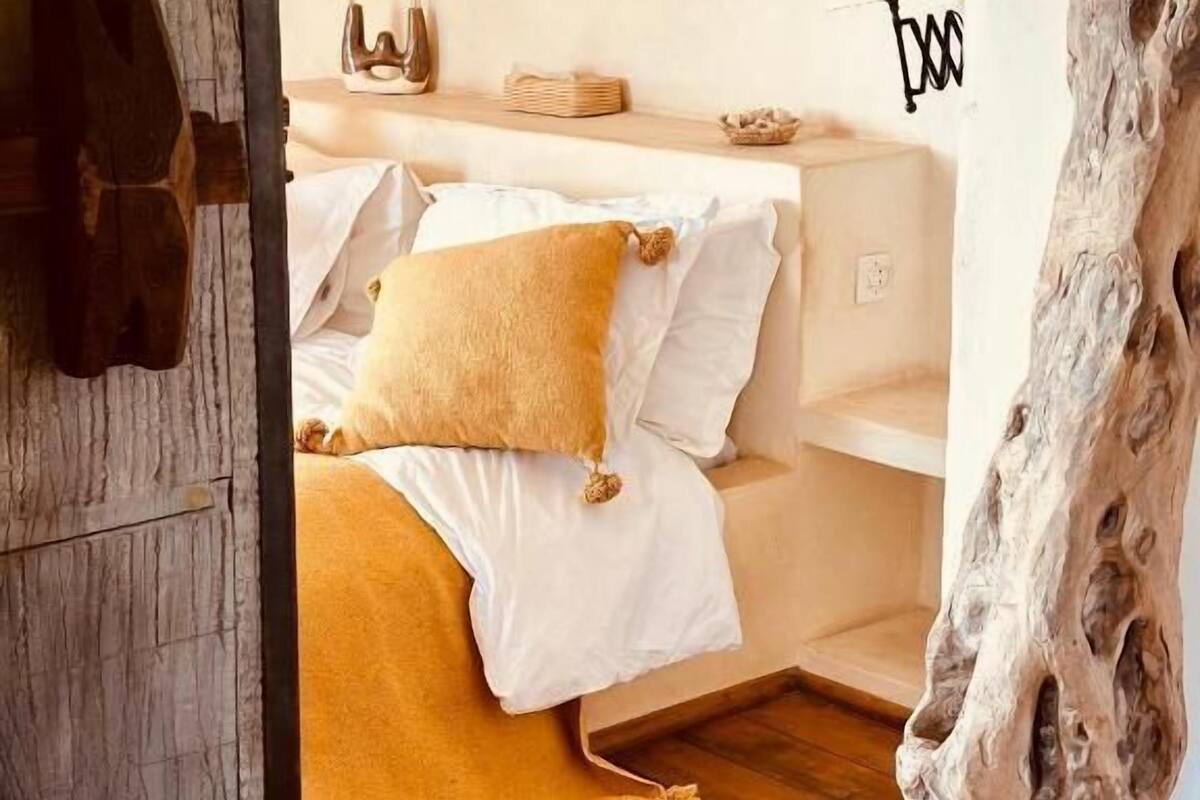 A bedroom with a wooden floor, a bed with white and mustard-colored bedding, a nightstand with a lamp, and a wall-mounted shelf with decorative items.