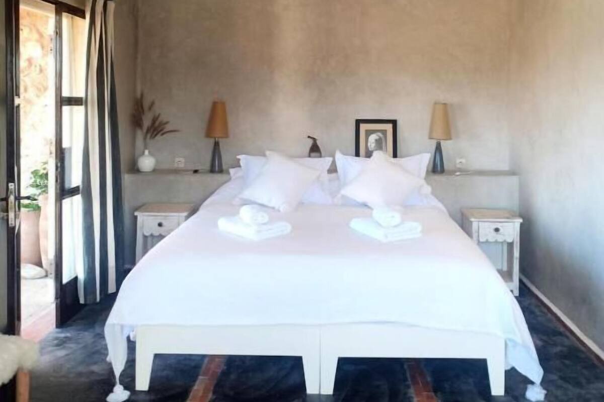 A large bed with white linens and pillows, two bedside tables with lamps, a small white nightstand, and a view of the outdoors through a sliding door.