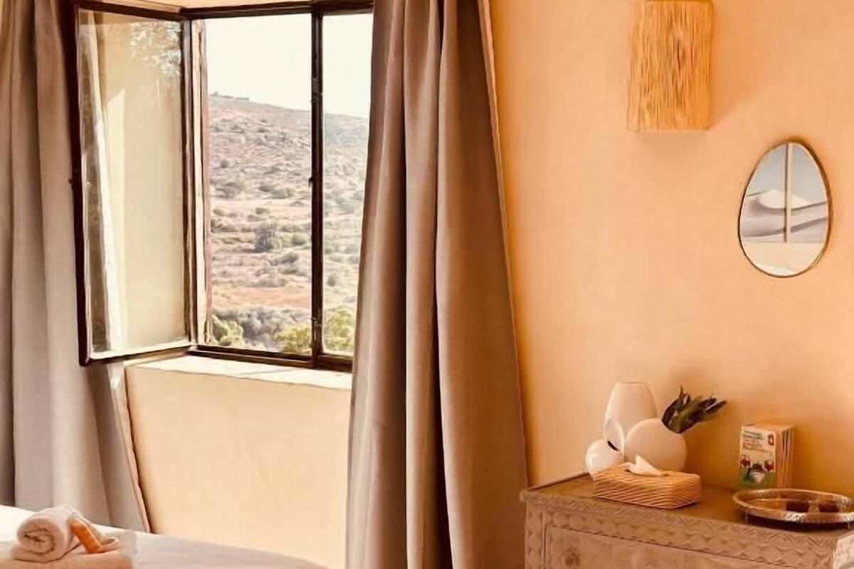 A bedroom with a bed, a nightstand, a window with curtains, and a view of a desert landscape.