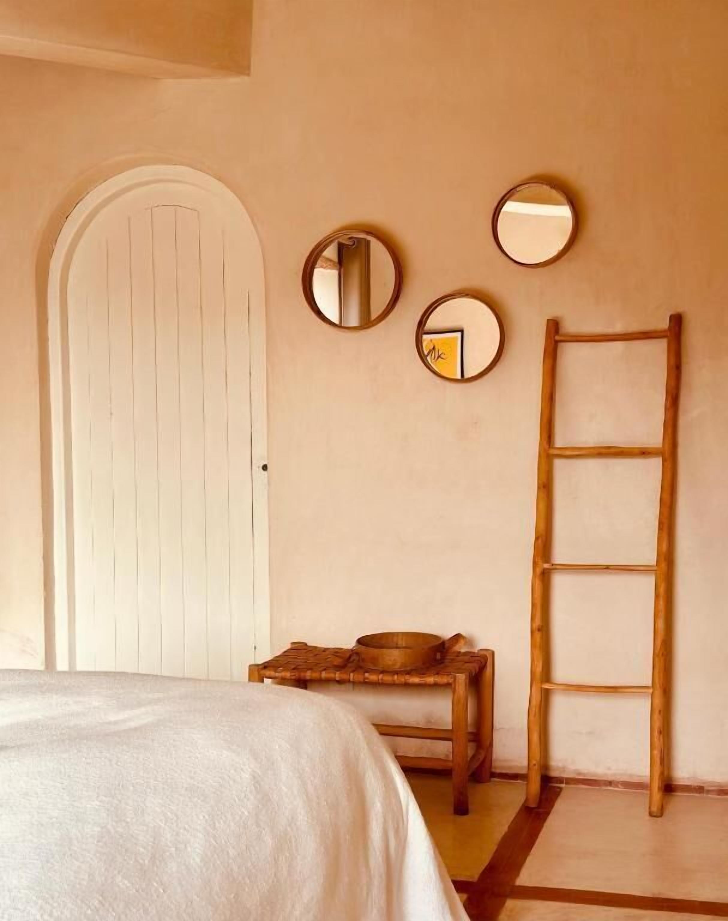 A bedroom with a bed, a wooden ladder, a small table with a bowl, and three round mirrors on the wall.
