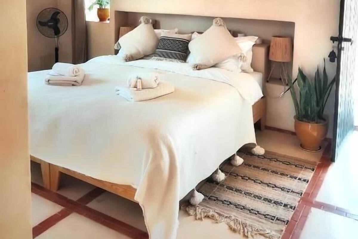 A neatly made bed with pillows and a folded towel, a standing fan, a potted plant, and a patterned rug.