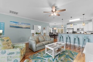 A spacious living room with a sofa, coffee table, and a view into the kitchen. - Walk to the Beach 3 BR Beach House with a Shared Pool! Pieradise (Garden City)