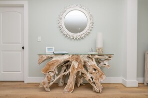 Ein Zimmer mit einer weißen Tür, einem runden Spiegel mit verziertem Rand, einem Glastisch mit natürlichem Holzfuß und einer weißen Vase auf dem Tisch.