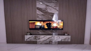 A modern living room with a flat-screen TV mounted on a marble console, flanked by vertical blinds. - Cozy 2 Bedroom Terrace Duplex + Wi-Fi,TV,24/7 Power In Ajah (Lekki/Ajah)