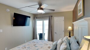 A bedroom with a bed, a ceiling fan, a TV mounted on the wall, a door, and a window with curtains. - Castle Beach 105 | Gulf-Front Retreat w/ Private Lanai + Pool (Fort Myers Beach)
