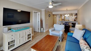 A living room with a TV, sofa, coffee table, and a dining area with chairs and a table. - Castle Beach 105 | Gulf-Front Retreat w/ Private Lanai + Pool (Fort Myers Beach)
