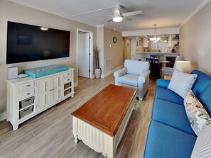 A living room with a TV, sofa, coffee table, and a dining area with chairs and a table. - Castle Beach 105 | Gulf-Front Retreat w/ Private Lanai + Pool (Fort Myers Beach)
