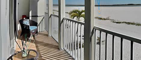 Varanda com vista para a praia, uma cadeira de vime, uma mesa e um corrimão.