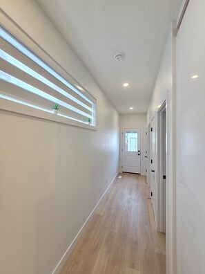 Ein Flur mit Holzboden, weißen Wänden, einem Fenster mit Jalousien, einer Tür am Ende und eingelassener Deckenbeleuchtung.