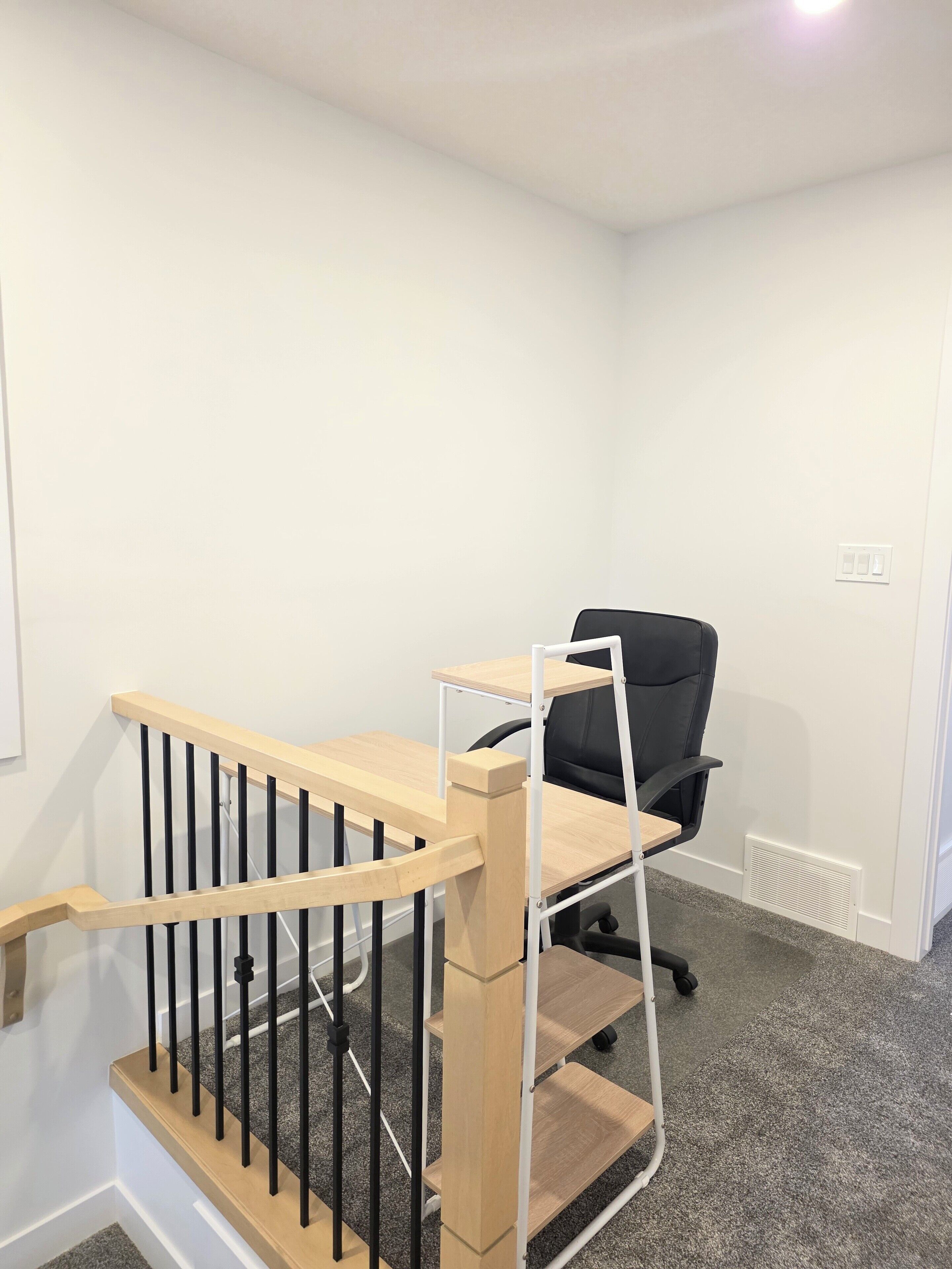 Eine Treppe mit Holzwah und schwarzen Spindeln, die zu einem Raum mit einem schwarzen Bürostuhl und einer weißen Wand führt.