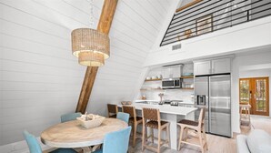A modern kitchen with white cabinetry, a stainless steel refrigerator, and a dining area with blue chairs and a wooden table. - A-Frame of Mind - Stylish Santa Rosa Beach Retreat | Walk to Beach (Santa Rosa Beach)