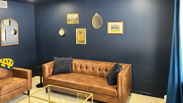 Een kamer met een donkerblauwe muur, een bruine leren bank, een kleine ronde tafel en een kroonluchter.