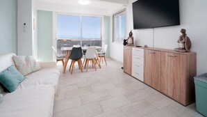 A modern living room with a sofa, a dining table with chairs, a TV mounted on the wall, and a view of the sea through large windows. - Light Filled Property Near Sea (Agde)