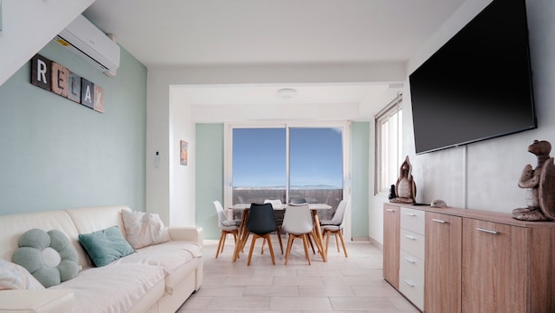 A modern living room with a sofa, dining table, chairs, and a TV mounted on the wall. - Light Filled Property Near Sea (Agde)