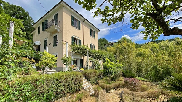 Ein zweistöckiges Gebäude mit Balkon, umgeben von üppiger Vegetation und einem Garten mit einem Steinweg.