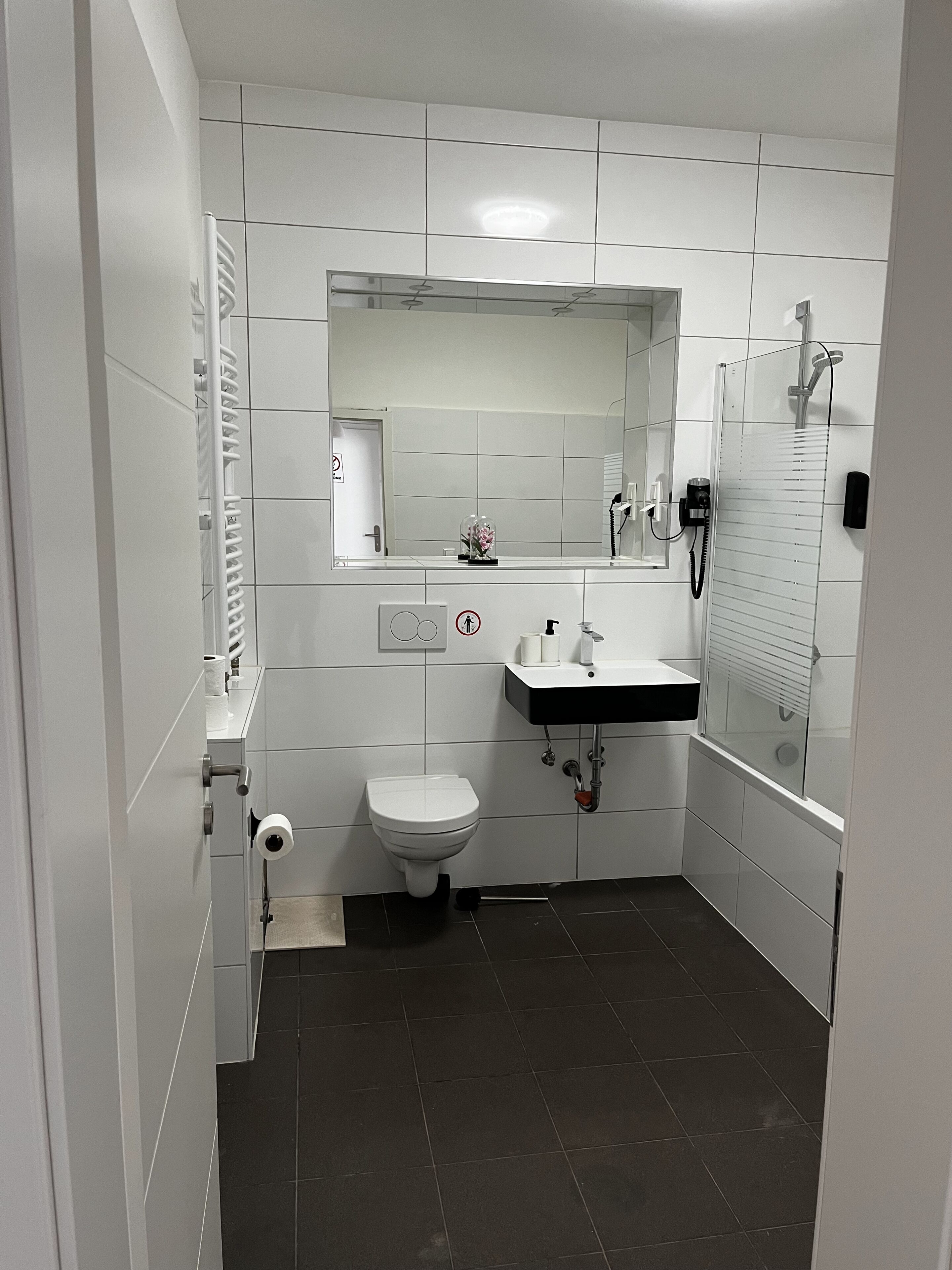A bathroom with white tiles, a toilet, a sink, a mirror, and a shower.
