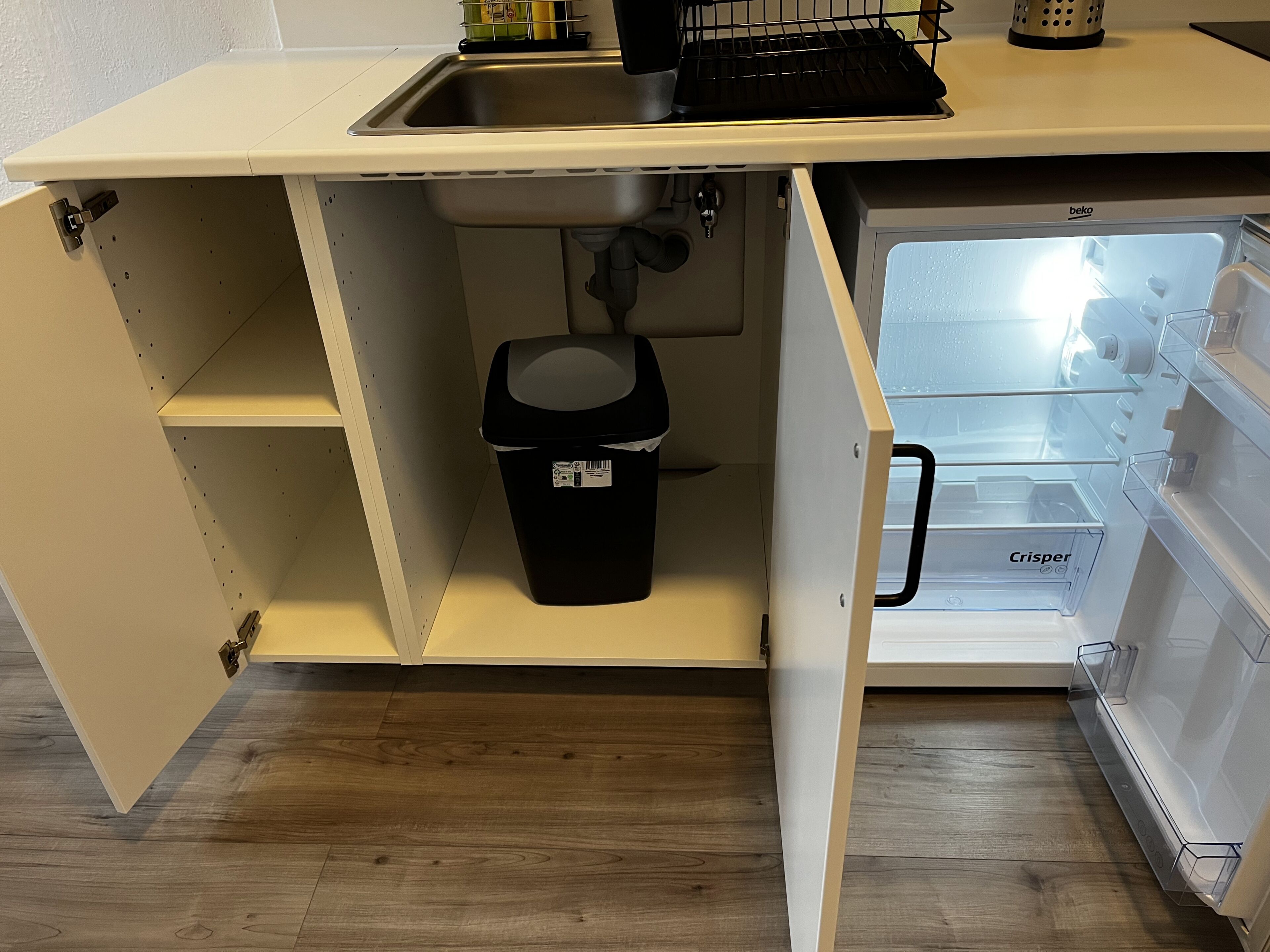 Una cocina con un lavaplatos empotrado, un cubo de basura y un refrigerador con puerta de vidrio.