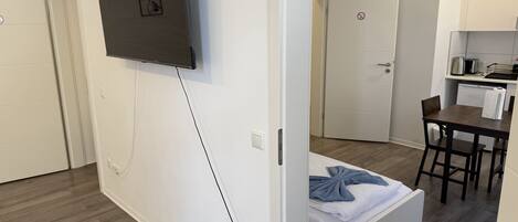 A modern, compact hotel room with a bed, a small table, a chair, a kitchenette, and a TV mounted on the wall.