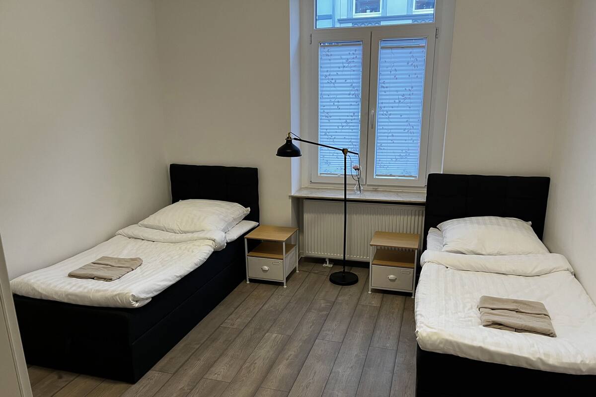 A small, clean room with two single beds, a nightstand, a floor lamp, and a window with blinds.