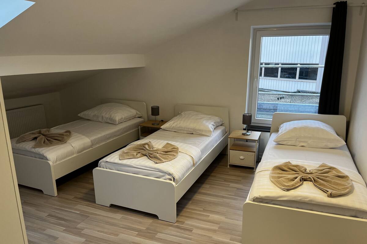 A room with three single beds, each with a bedside table and lamp. The room has a sloped ceiling and a window with curtains.
