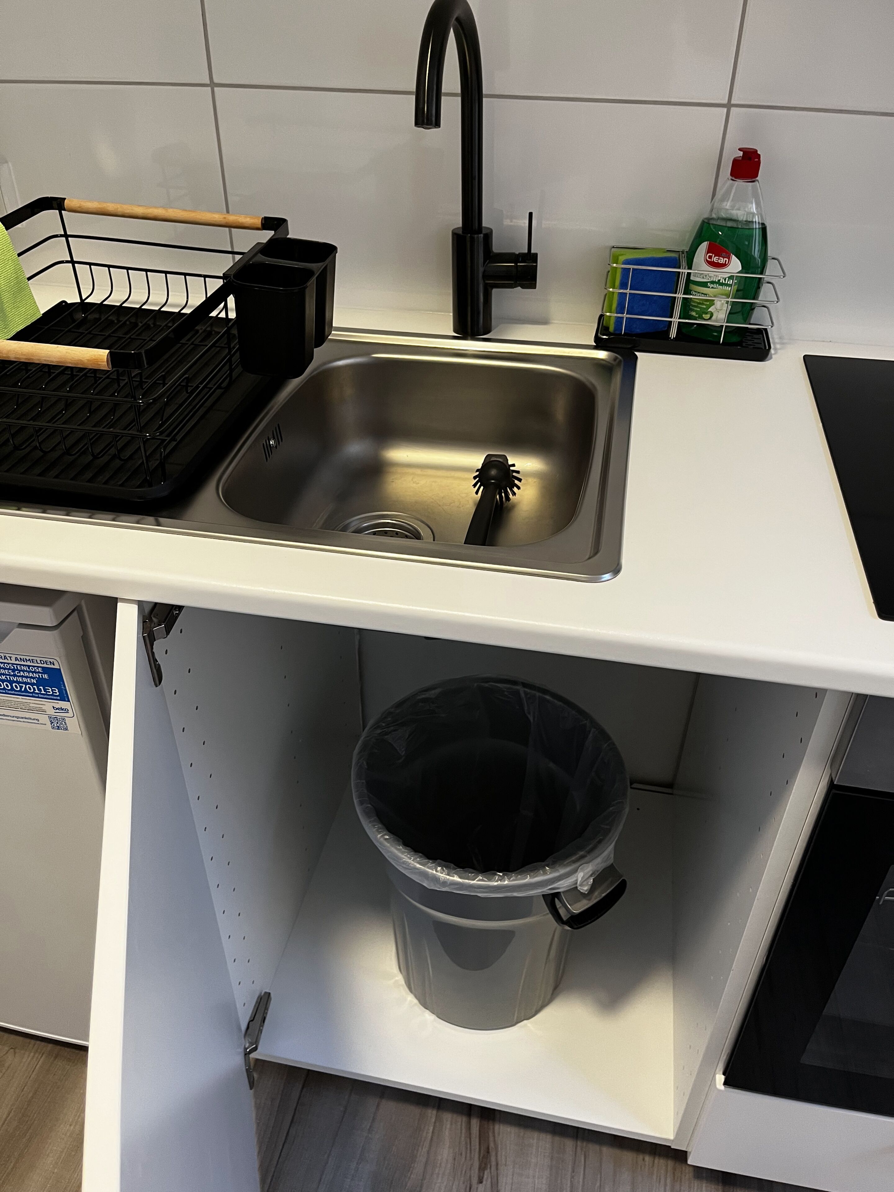 Una cocina con un lavamanos de acero inoxidable, un escurreplatos negro, una botella de detergente para trastes verde y un cubo de basura gris.