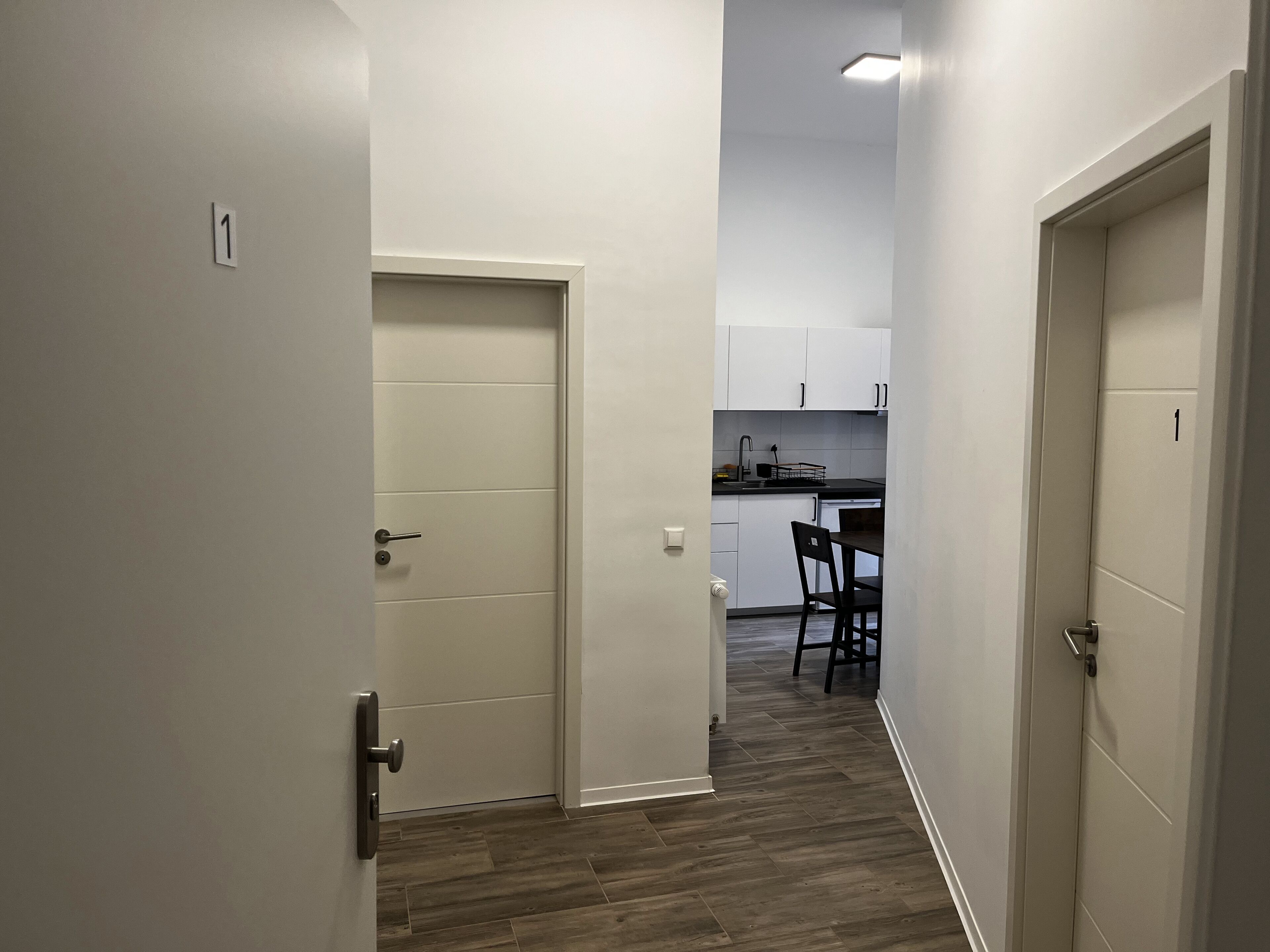 A narrow hallway with a wooden floor, leading to a kitchen area with white cabinets and a dining table with chairs.