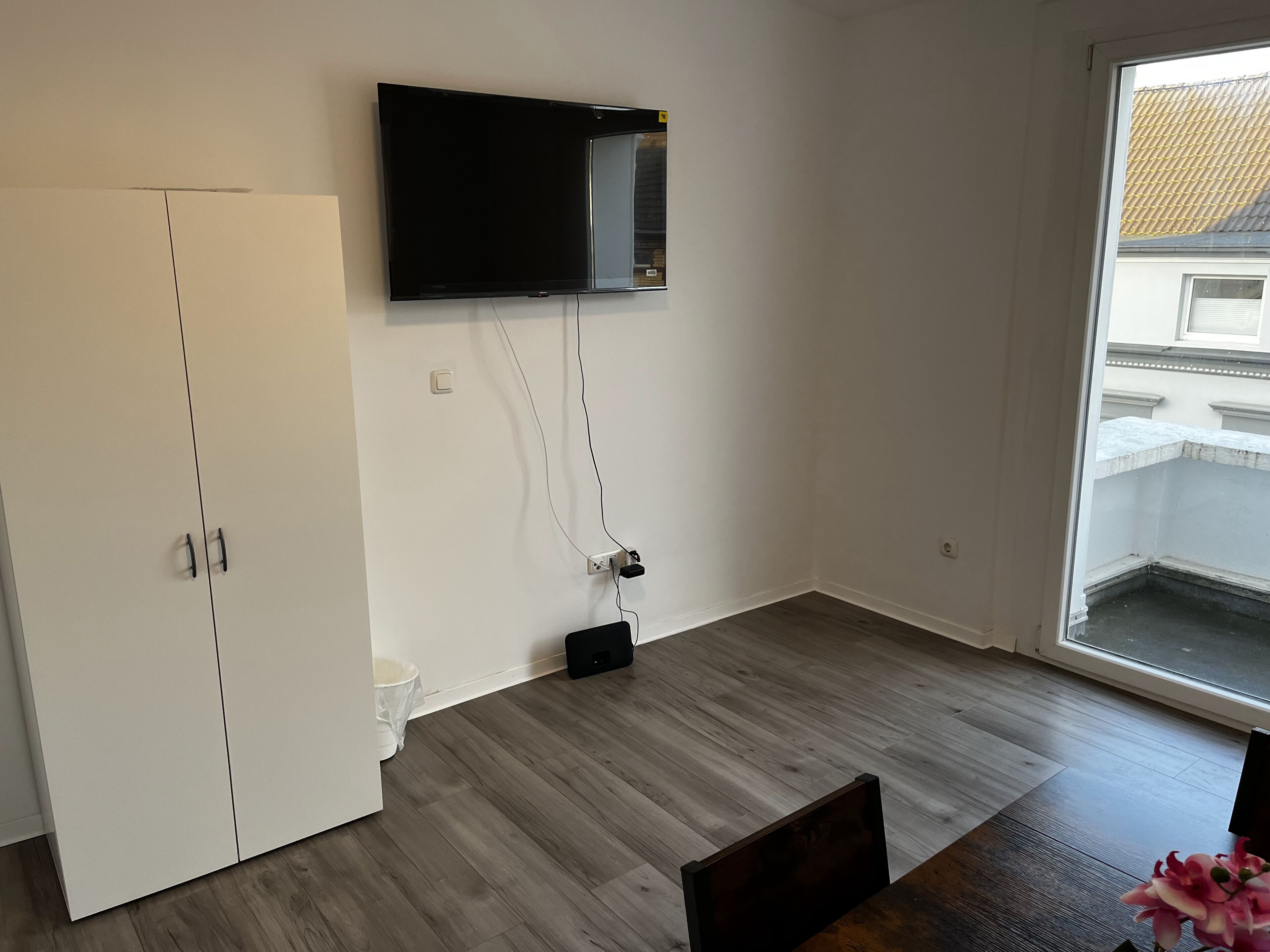 A room with a flat-screen TV mounted on the wall, a white wardrobe, and a dining table with chairs.
